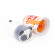 Gray and white panda Pandaroll plush toy lying next to a Cup Noodles-themed plush cup tipped on its side, revealing orange noodle and narutomaki toppings inside, on a white background
