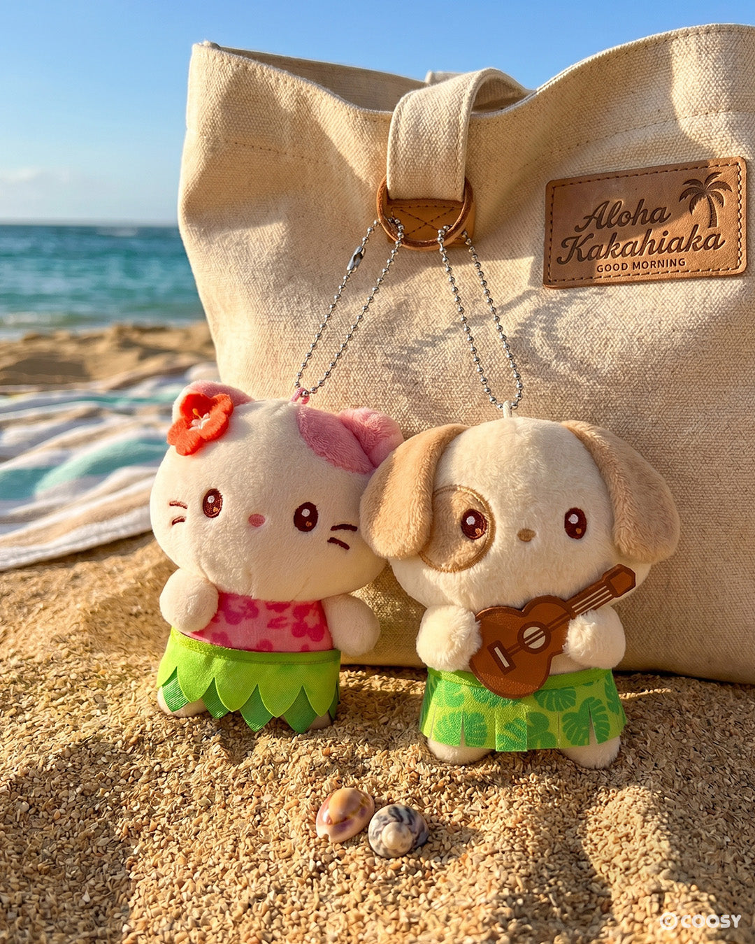 Two Miniz Hawaii Collection plush keychains — Kittiroll in a pink floral top and green hula skirt with a hibiscus flower, and Puppiroll holding a ukulele in a green tropical skirt — clipped to a canvas tote bag on a sunny beach.
