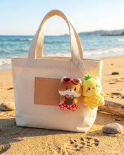 Two Miniz by Anirollz Hawaii Collection plush keychains — Foxiroll in red floral swim trunks and sunglasses holding a coconut drink, and  yellow Chickiroll dressed as a pineapple — clipped to a canvas tote bag on a sunny beach.